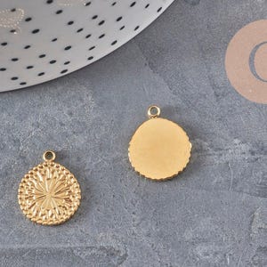 Peut inclure: Deux breloques rondes en métal doré avec de petites boucles sur le dessus. L'une des breloques présente un motif floral ou en forme de soleil en relief sur sa face avant, tandis que l'autre affiche une surface dorée lisse, plate et unie.
