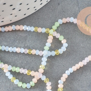 May include: Pastel-coloured beaded strands in pink, blue, yellow, and green. The small, faceted beads create a sparkling effect. The beads are arranged on a grey surface, with a white and black dotted ceramic bowl in the background.