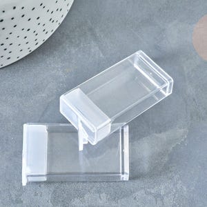 May include: Two rectangular, clear plastic storage containers with hinged lids. Each container features a smooth, transparent body and a white, opaque latch mechanism on one end, designed for organising small items in a compact, modular fashion.
