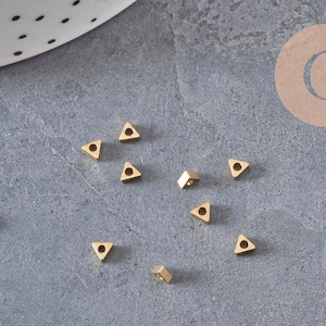 May include: A collection of ten small, gold-colored metal beads shaped like equilateral triangles. Each bead features a centered circular hole, providing a uniform appearance. The beads are scattered across the surface, displaying a metallic finish.