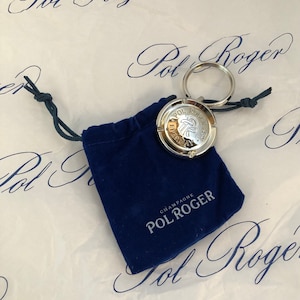 Puede incluir: Un llavero plateado con un dije redondo que presenta el logotipo de Pol Roger y las palabras "Everyday" y "Champagne Pol Roger". El llavero está en una bolsa de terciopelo azul.