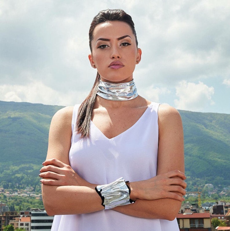 Puede incluir: Una mujer con el pelo largo y oscuro lleva una blusa blanca sin mangas y un collar de gargantilla plateado. Tambi&eacute;n lleva una pulsera plateada en la mu&ntilde;eca. Est&aacute; de pie frente a una cordillera.