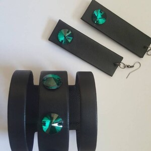May include: A set of black leather jewelry with green crystal accents. The set includes a bracelet with three bands and a pair of earrings.