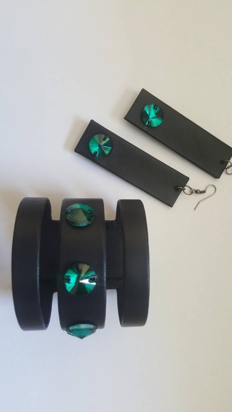 May include: A set of black leather jewelry with green crystal accents. The set includes a bracelet with three bands and a pair of earrings.