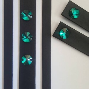 May include: Black leather straps with three emerald green rhinestones attached. The straps are cut into different lengths.