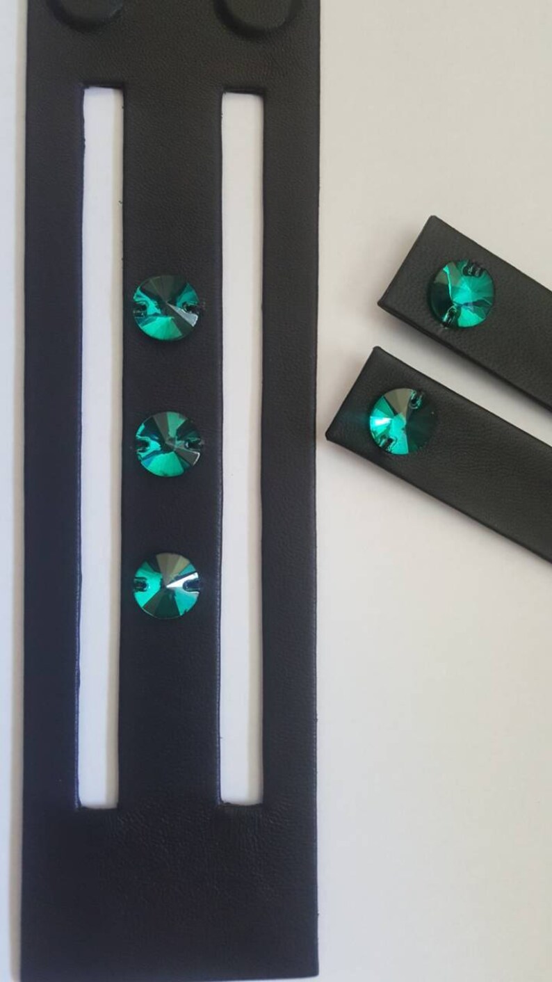 May include: Black leather straps with three emerald green rhinestones attached. The straps are cut into different lengths.