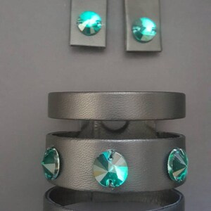 May include: A set of black leather jewelry featuring a layered bracelet with three bands and three teal rhinestones and a pair of matching earrings with teal rhinestones.