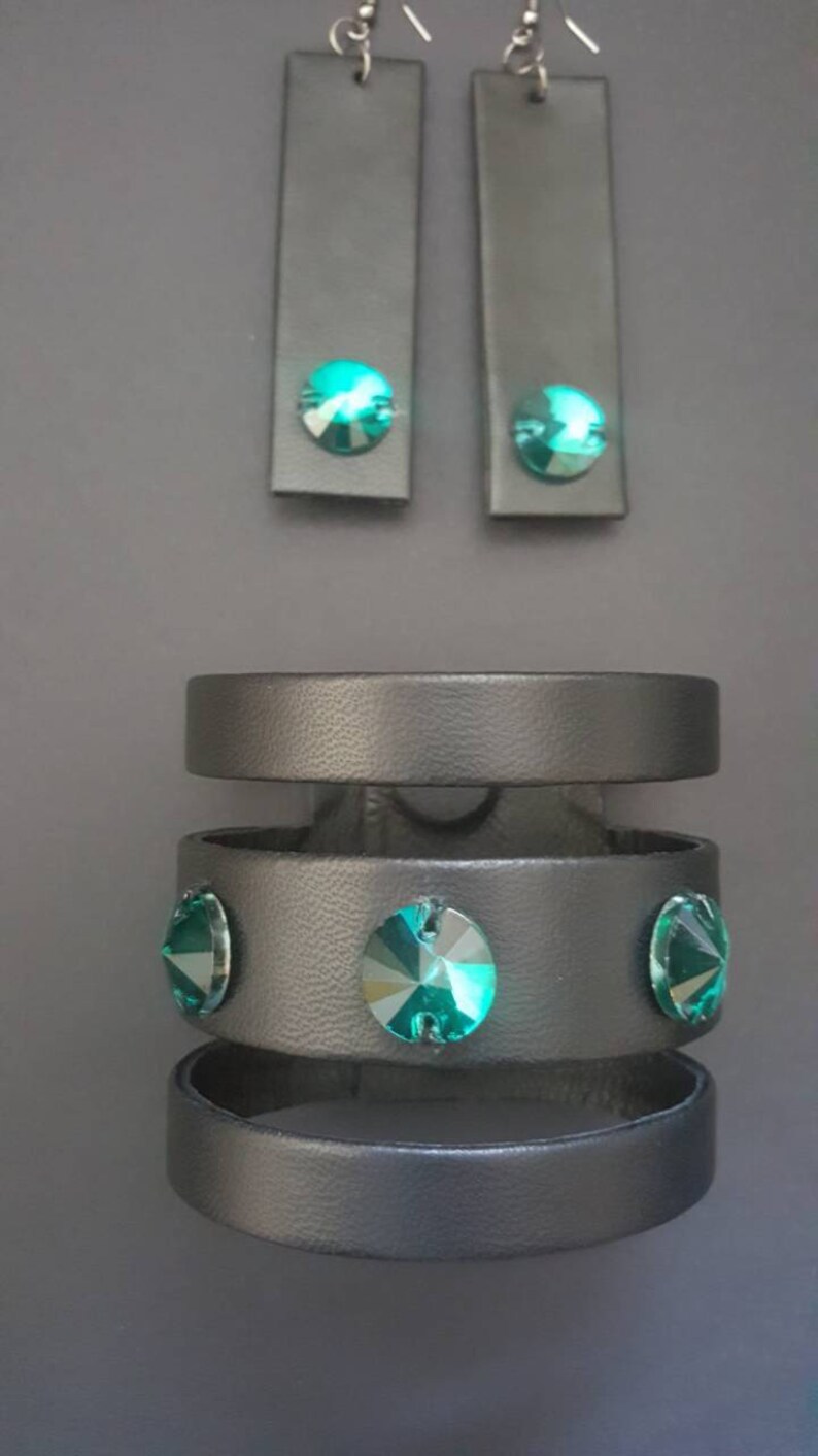 May include: A set of black leather jewelry featuring a layered bracelet with three bands and three teal rhinestones and a pair of matching earrings with teal rhinestones.