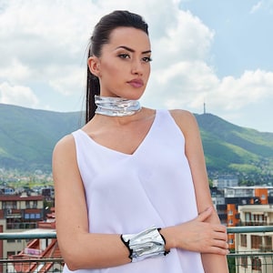 Puede incluir: Una mujer con un top blanco sin mangas, un collar de gargantilla plateado y una pulsera plateada. Est&aacute; de pie frente a un horizonte urbano con una monta&ntilde;a en el fondo.