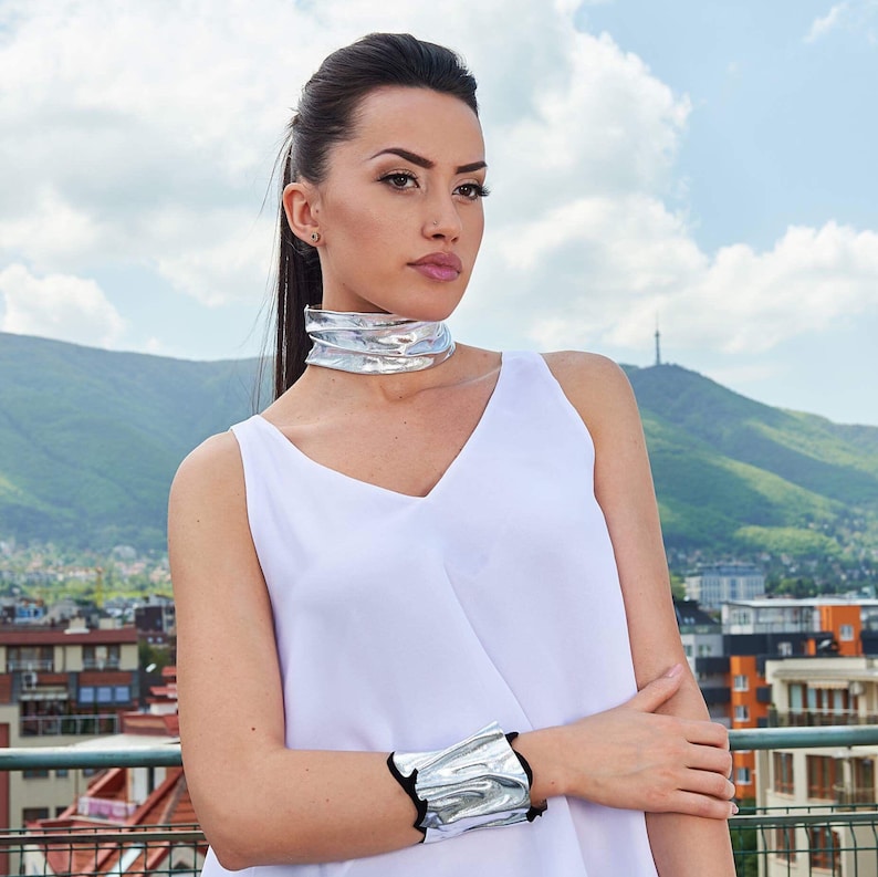 Puede incluir: Una mujer con un top blanco sin mangas, un collar de gargantilla plateado y una pulsera plateada. Est&aacute; de pie frente a un horizonte urbano con una monta&ntilde;a en el fondo.