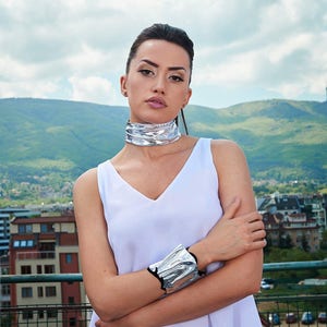 Puede incluir: Una mujer con un top blanco sin mangas, un collar de gargantilla plateado y una pulsera plateada. Est&aacute; de pie frente a una cordillera.