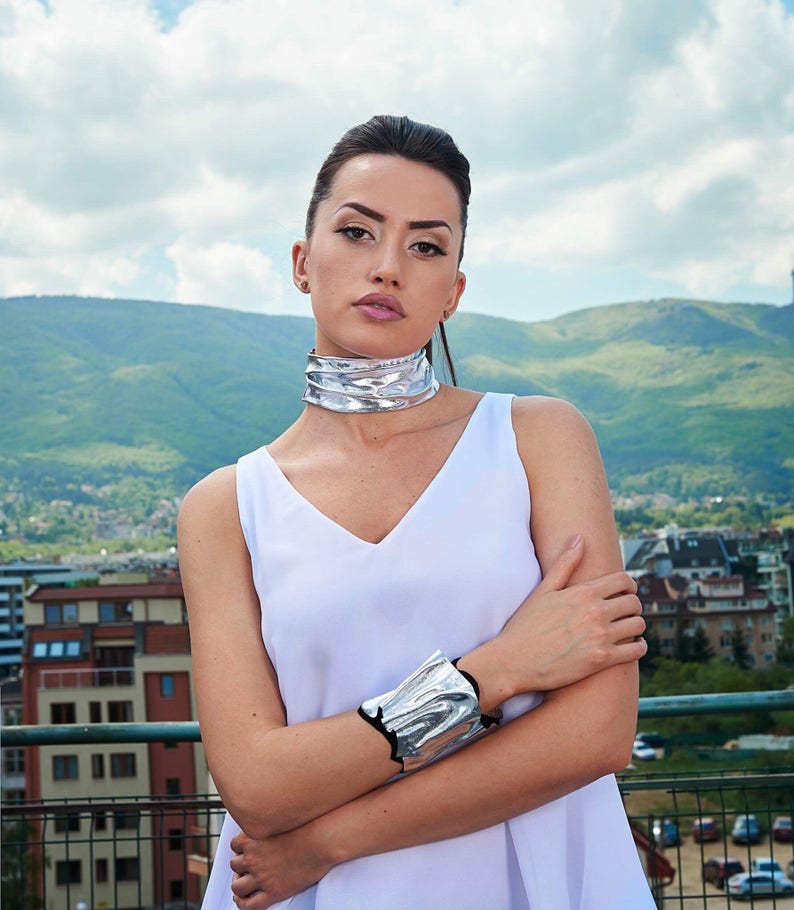 Puede incluir: Una mujer con un top blanco sin mangas, un collar de gargantilla plateado y una pulsera plateada. Est&aacute; de pie frente a una cordillera.