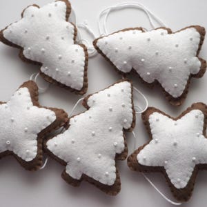 May include: A collection of handmade felt Christmas ornaments in the shapes of stars and Christmas trees. The ornaments are white with brown edges and small white bead embellishments. Each ornament has a white string for hanging.