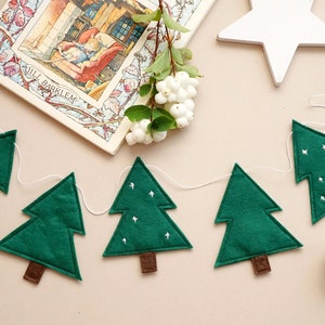 May include: A green felt Christmas tree garland with white felt snowflakes. The garland is strung on white twine and has brown felt tree trunks. The garland is hanging on a light brown background.
