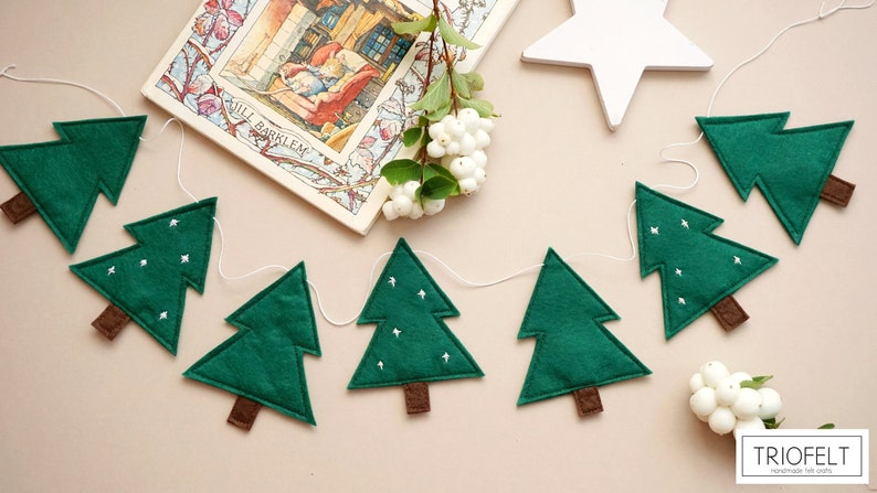 May include: A green felt Christmas tree garland with white felt snowflakes. The garland is strung on white twine and has brown felt tree trunks. The garland is hanging on a light brown background.