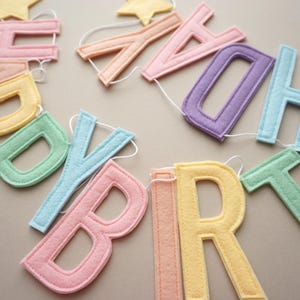 May include: A felt letter garland spelling out HAPPY BIRTHDAY in pastel colors including pink, yellow, green, blue, and purple. The individual letters are connected by a thin white string and accented with small yellow felt stars at the top.