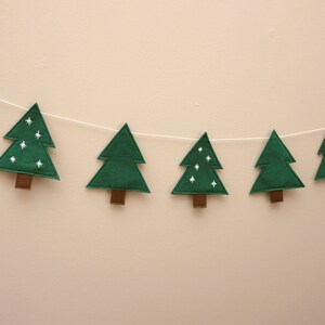 May include: A green felt Christmas tree garland with white embroidered stars. The garland is strung on a white string and hangs against a white wall.