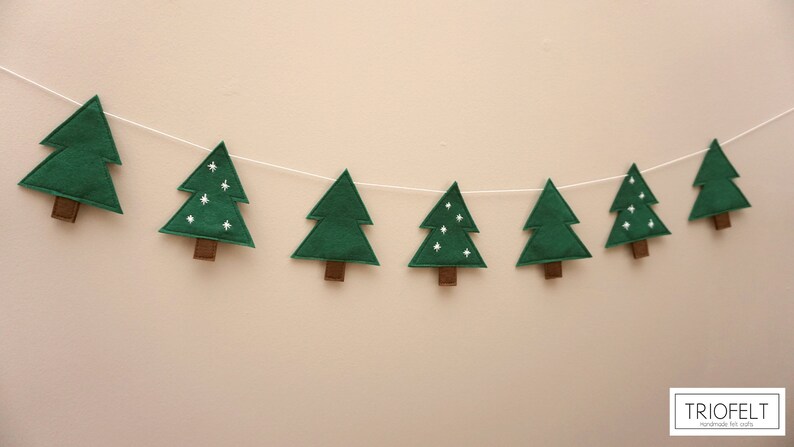 May include: A green felt Christmas tree garland with white embroidered stars. The garland is strung on a white string and hangs against a white wall.