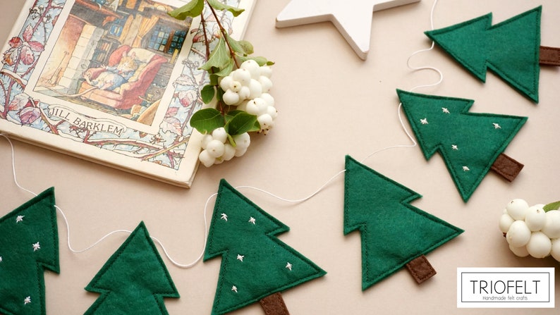 May include: A green felt Christmas tree garland with white felt stars, brown felt trunks, and white string. The garland is hanging on a light pink surface. The text "TRIOFELT handmade felt crafts" is visible in the bottom right corner of the image.