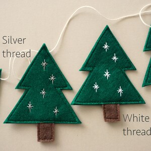 May include: A garland of green felt Christmas trees with white and silver thread embroidery. The trees are strung together with white thread. The text "Silver thread" and "White thread" is written on the image.