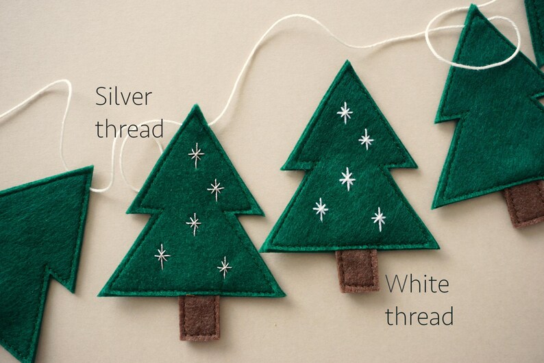 May include: A garland of green felt Christmas trees with white and silver thread embroidery. The trees are strung together with white thread. The text "Silver thread" and "White thread" is written on the image.