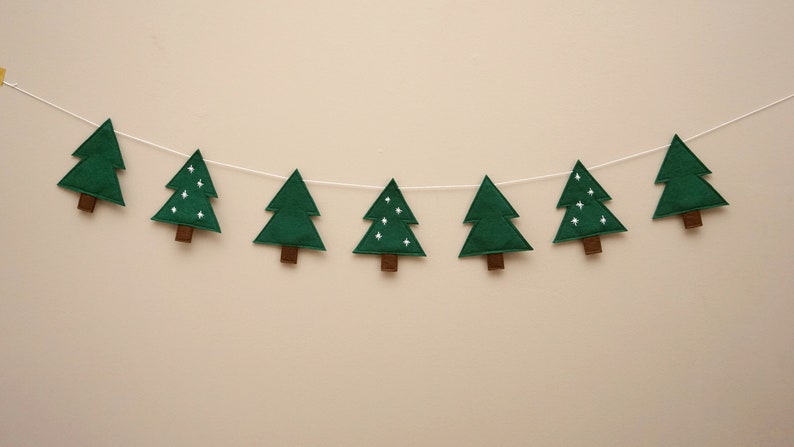 May include: A green felt Christmas tree garland with white stars. The garland is strung on a white string and has eight trees.