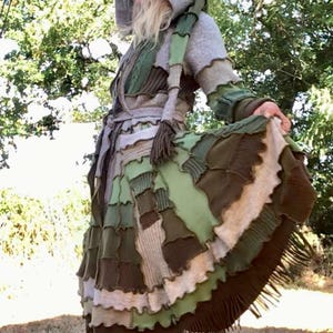 May include: A hooded coat featuring a patchwork design of various green, brown, and grey fabric textures. The garment includes a tie waist, long sleeves, and a flared skirt section with raw-edge seams and a fringe trim along the bottom hem.