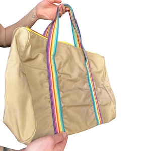 Pode incluir: Bolsa tote bege com alça e detalhes listrados em arco-íris. A bolsa tem um zíper amarelo e é segurada pelas mãos de uma pessoa. A bolsa é feita de um material liso, possivelmente nylon.