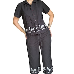 May include: Black two-piece outfit featuring a short-sleeved button-down shirt and cropped pants. The shirt and pants have white embroidered palm tree designs along the edges. The pants have a drawstring waist.
