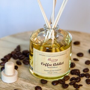 Op de afbeelding: Een heldere glazen fles met een bruin etiket waarop "bloomey handmade" en "Coffee Addict Reed Diffuser Handmade Air Freshener 100ml/3.34 oz." staat. De fles is gevuld met een lichtbruine vloeistof en heeft houten stokjes die uit de bovenkant steken. De fles is omringd door koffiebonen op een houten oppervlak.