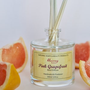Può includere: Una bottiglia di vetro trasparente con un'etichetta di legno che recita "bloomey handmade Pink Grapefruit Reed Diffuser Handmade Air Freshener 100ml/3.34 oz.". La bottiglia è piena di un liquido trasparente e ha bastoncini di legno che sporgono. Ci sono fette di pompelmo rosa intorno alla bottiglia.