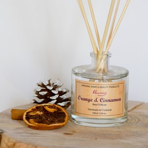 May include: A clear glass bottle with a brown label that reads "bloomey handmade" and "Orange & Cinnamon Reed Diffuser Handmade Air Freshener 100ml/3.34 oz." The bottle is filled with a clear liquid and has wooden sticks sticking out of the top. The bottle is sitting on a wooden surface with a cinnamon stick and a dried orange slice.