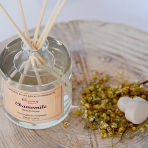 Può includere: Una bottiglia di vetro trasparente con un'etichetta marrone che recita "Chamomile Reed Diffuser Handmade Air Freshener 100ml/3.34 fl. oz.". La bottiglia è piena di un liquido trasparente e ha bastoncini di legno inseriti al suo interno. La bottiglia è appoggiata su una superficie di legno con fiori di camomilla secchi sparsi intorno.