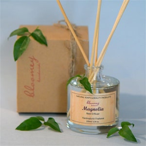 Può includere: Una bottiglia di vetro trasparente con un'etichetta marrone su cui è scritto "bloomey handmade Magnolia Reed Diffuser Handmade Air Freshener 100ml/3.34 oz.". La bottiglia è piena di un liquido trasparente e ha bastoncini di legno inseriti al suo interno. La bottiglia è appoggiata su una superficie marrone chiaro con foglie verdi sparse intorno.