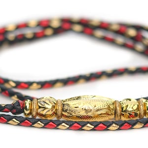 May include: A braided cord necklace featuring a central gold-toned metal bead with intricate etched patterns, flanked by two smaller matching gold-toned beads. The cord is woven from black, red, and metallic gold strands in a repeating pattern.