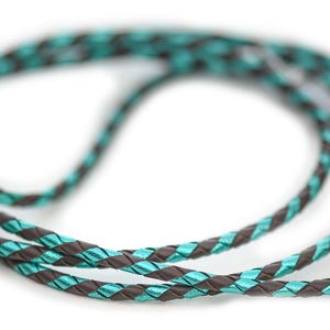 May include: A braided cord featuring a repeating pattern of dark brown and vibrant turquoise strands. The cord is coiled into a circular shape, showcasing the intricate woven texture of the material throughout the entire length of the flexible cord.