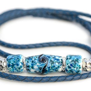 May include: A blue leather necklace with a braided design. The necklace features three decorative beads: two square beads with a speckled blue pattern and a central cylindrical bead with a swirl design. Silver-colored metal beads separate the decorative beads.