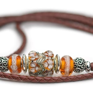 May include: A brown braided leather cord bracelet featuring a central glass bead with a white and green floral pattern, flanked by two orange glass beads, two silver-toned metal spacers, and two ornate silver-toned metal beads on each side.