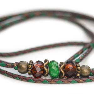 May include: A braided cord necklace featuring a mix of brown and green strands. The center is adorned with two faceted amber-colored beads, one central faceted green bead, and several metallic bronze-toned spacer beads and wavy metal accents.