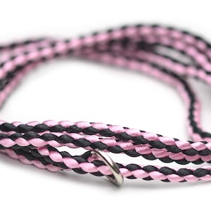 May include: A braided cord necklace featuring alternating strands of black and light pink satin-like material. The cord is coiled in a circular shape and includes a single, small, circular silver-toned metal ring attached to one section of the braid.