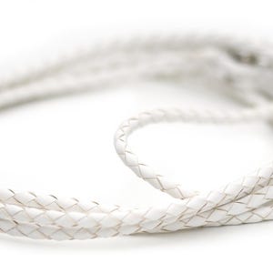 May include: A white, braided leather cord, likely for a necklace or other jewelry. The cord is tightly woven and appears to be made of multiple strands. The cord is coiled and lies on a white background.