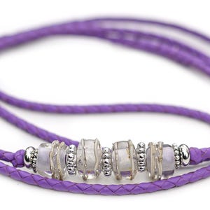 May include: A braided purple leather necklace with a central beaded design. The necklace features clear, rectangular beads interspersed with silver-colored metal beads. The necklace is a delicate piece of jewelry.