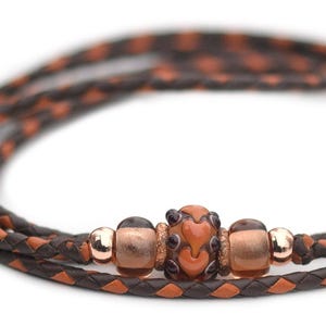 May include: A brown and orange leather cord with a woven pattern and a copper and orange glass bead.