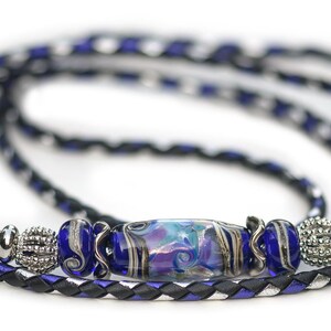 May include: A braided cord necklace featuring a central glass bead with blue, purple, and white swirls, flanked by smaller blue glass beads, silver-toned textured metal spacers, and a long, thin cord woven with black, blue, and silver metallic strands.