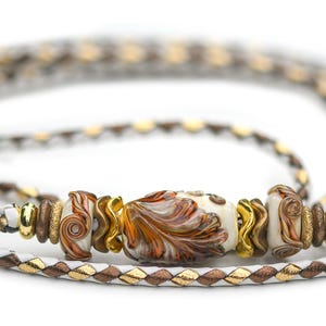 May include: A multi-strand wrap bracelet featuring a braided cord in white, brown, and metallic gold. The center is adorned with a series of decorative glass beads in white, orange, and brown swirls, accented by small gold-toned metal spacers.