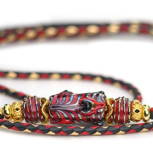 May include: A braided cord bracelet featuring a central focal bead with red, black, and white swirling patterns, flanked by smaller red and black striped beads and gold-toned metal spacers. The cord is woven with black, red, and gold-toned strands.