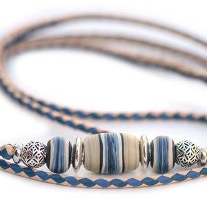 May include: A braided leather necklace with blue and tan tones. The necklace features a central, oblong bead with blue and tan stripes, flanked by two blue and white striped beads and two silver filigree beads. The necklace is approximately 30 inches long.