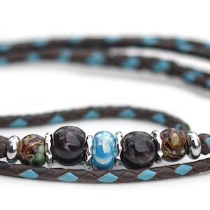 May include: A brown and turquoise leather wrap bracelet with decorative beads. The bracelet features a braided design with turquoise accents and a series of dark and light-colored beads. The beads are separated by silver-colored metal rings.