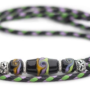 May include: A black and green braided leather cord with a silver and glass bead accent. The bead is black with swirls of yellow, orange, and purple.