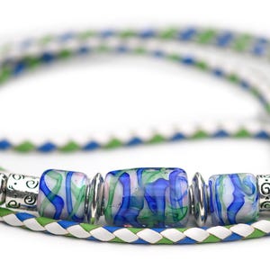 May include: A braided cord bracelet featuring white, blue, and green strands. The center is adorned with three glass beads displaying a marbled blue and green pattern, separated by silver-toned metal spacers with etched decorative details.